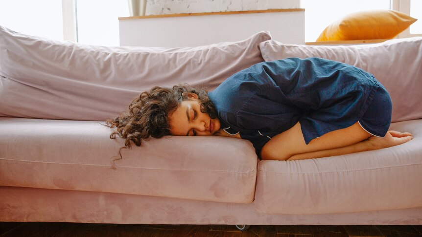 一名身穿蓝色睡衣的女性蜷缩在一个球，在粉红色的沙发上痛苦” class=